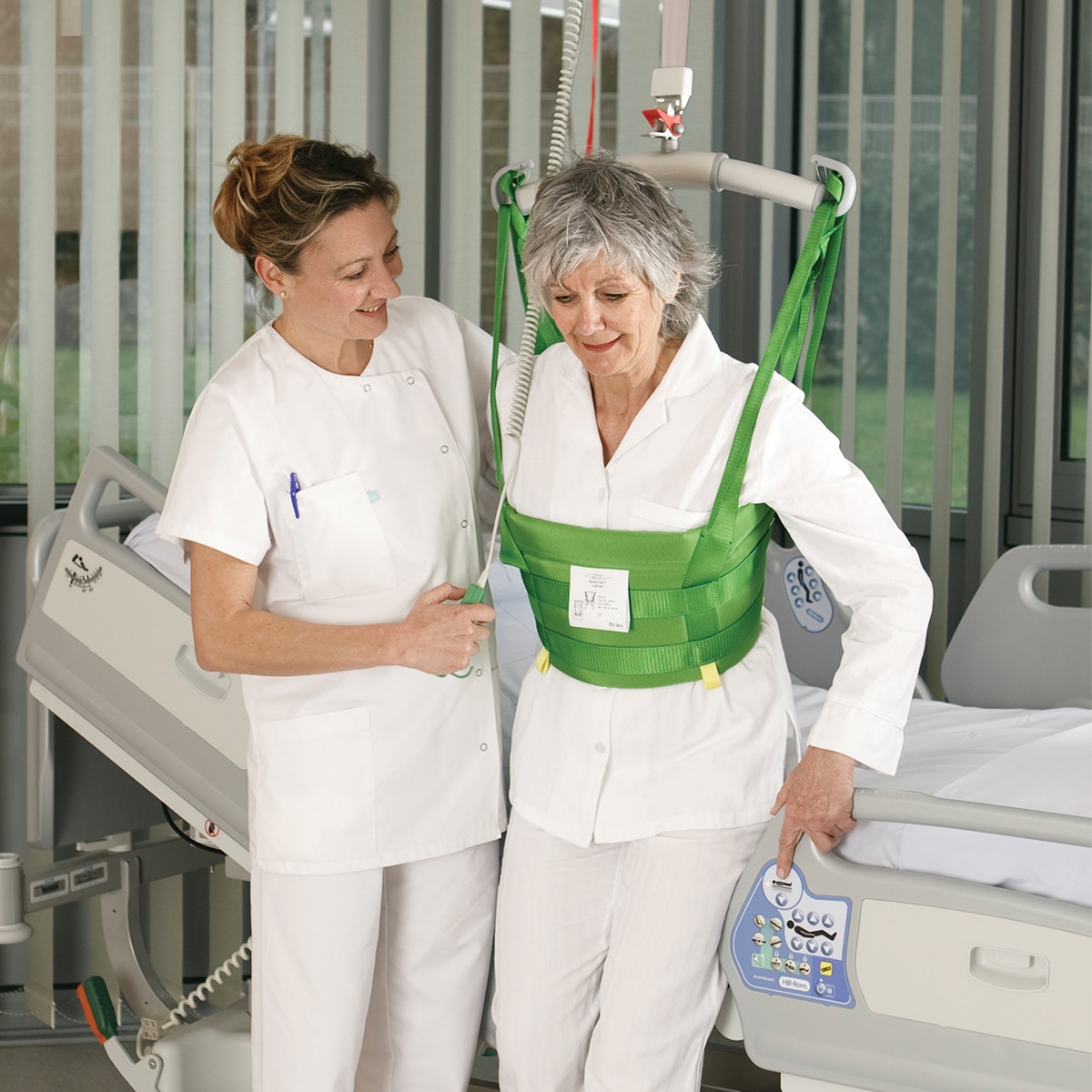 A clinician helps a patient walk in a hospital room with a Hillrom overhead lift and MasterVest